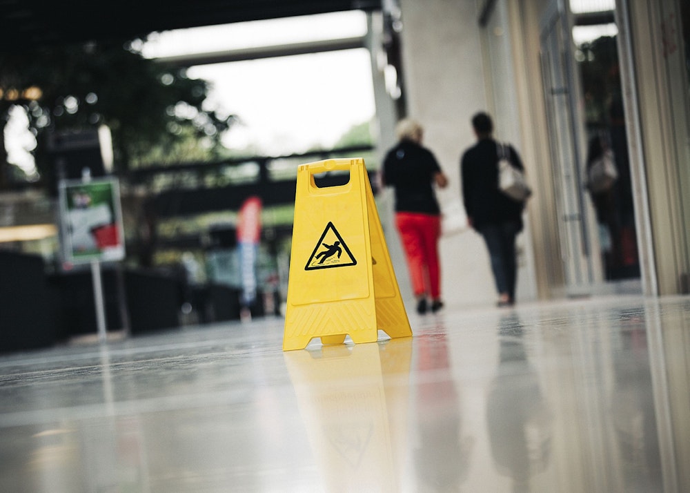 A wet floor sign is seen in the foreground while people walk in front of an office building in the background.