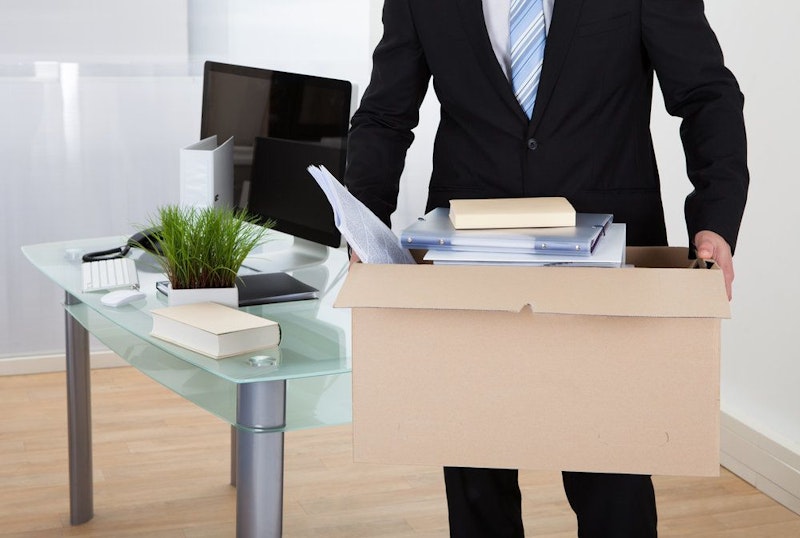 A man in a suit carries a box of personal belongings away from his desk.