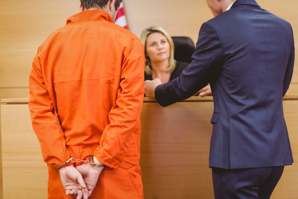 Man wearing an orange jumpsuit accompanied by his lawyer, speaking to a judge.