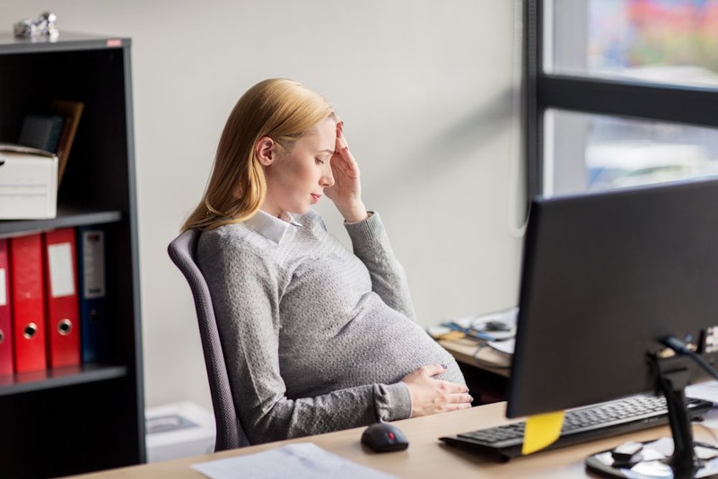 Pregnant woman sitting at a desk with her hand to her head.