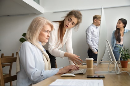 Older woman at keyboard scowling as younger woman leans over her workspace