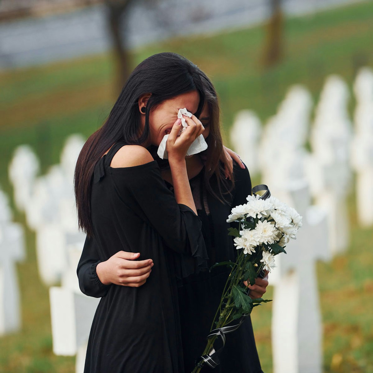 Two grieving women embracing in a cemetery, one holding white flowers and wiping tears with a tissue.