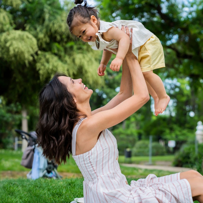 Young mother sitting on blanket outdoors, holding child up in the air
