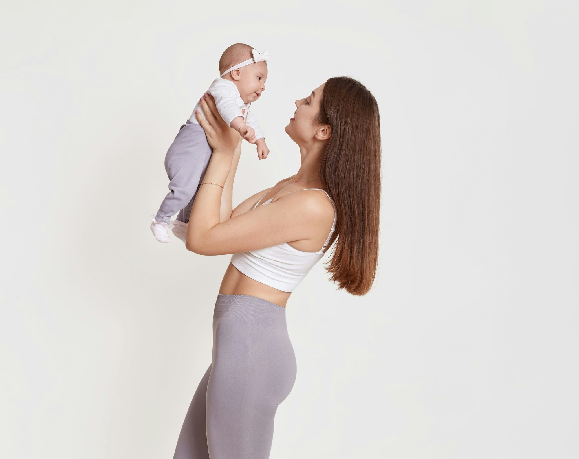 Slim woman wearing white bra and yoga pants lifting up baby