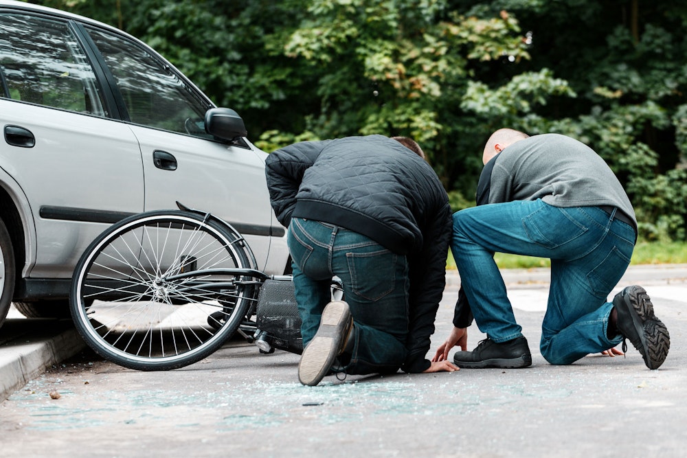 Two people kneeling beside a fallen bicycle next to a car after a roadway collision