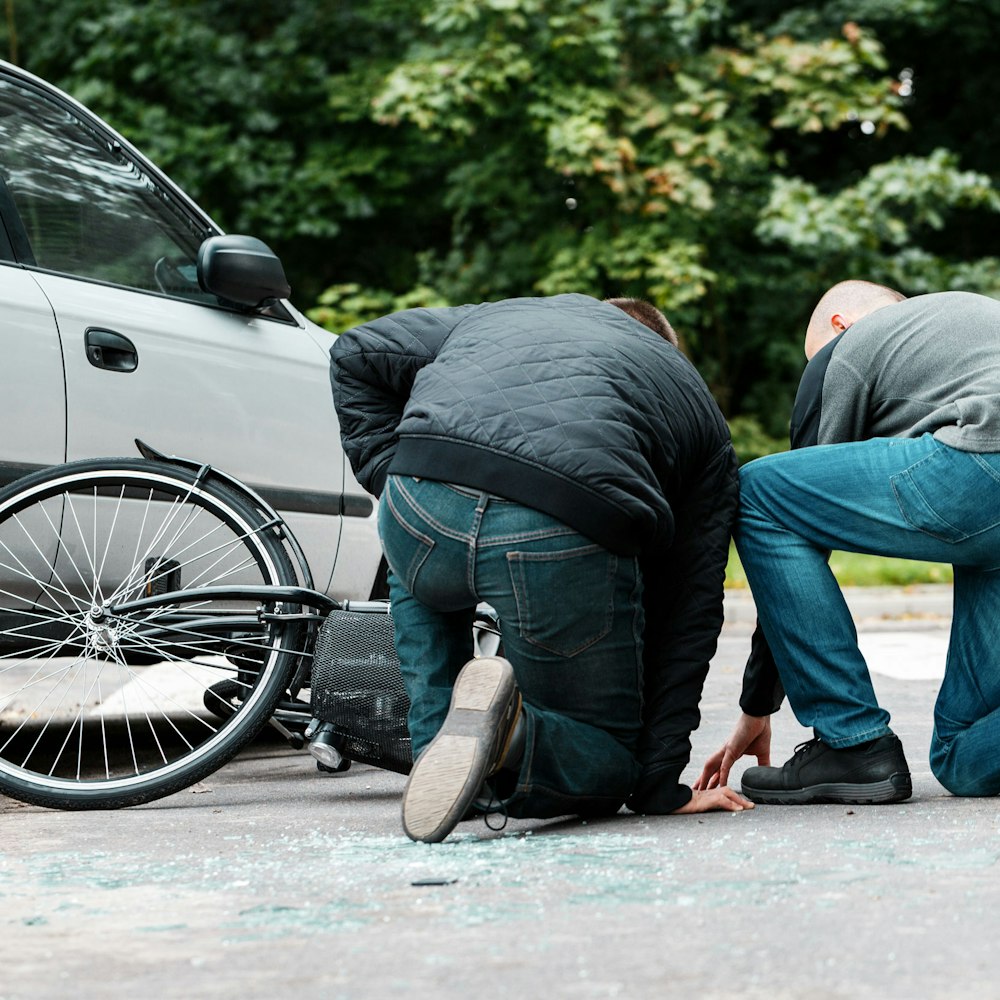Two people kneeling beside a fallen bicycle next to a car after a roadway collision