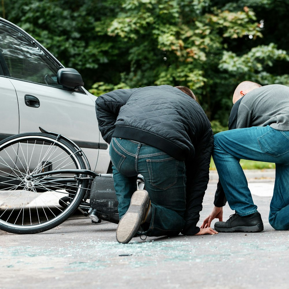Two people kneeling beside a fallen bicycle next to a car after a roadway collision